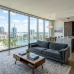 Bright, modern apartment with floor-to-ceiling windows overlooking the Orlando skyline and Lake Eola. Features a gray velvet sofa, wooden floors, and a luxury open kitchen with a marble island.